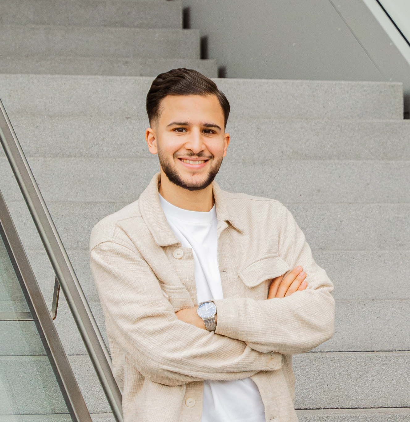 Young man with neatly styled dark hair and beard smiling with arms crossed, standing in front of gray stairs and glass railing.