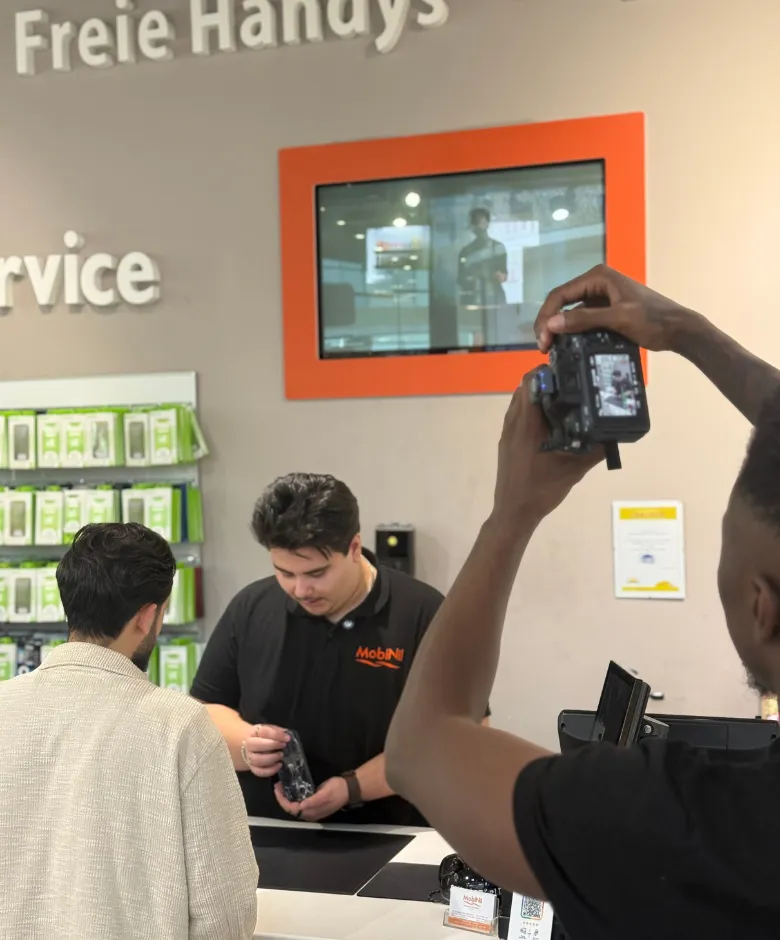 A camera operator filming a man with dark hair and mustache at a service counter holding a phone case, while a customer in a light-colored jacket watches.