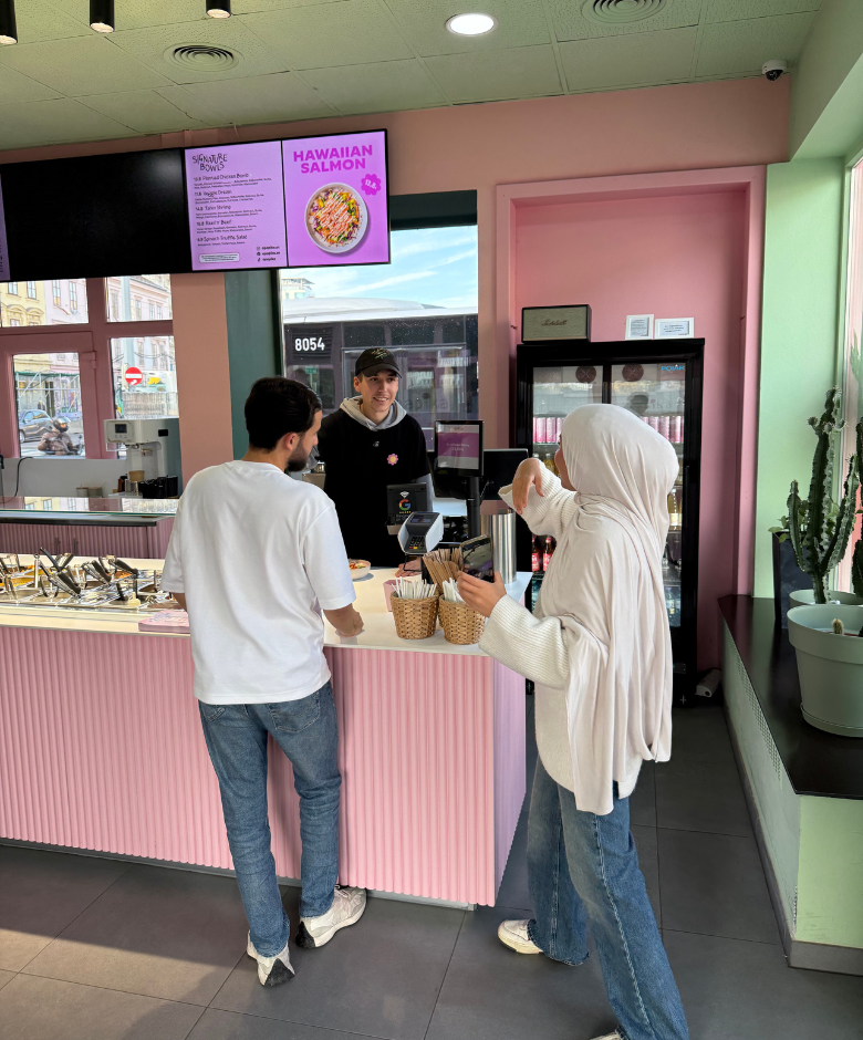 A woman wearing a hijab and a man stand at a pink counter ordering food from a smiling employee in a casual eatery with a menu screen overhead.