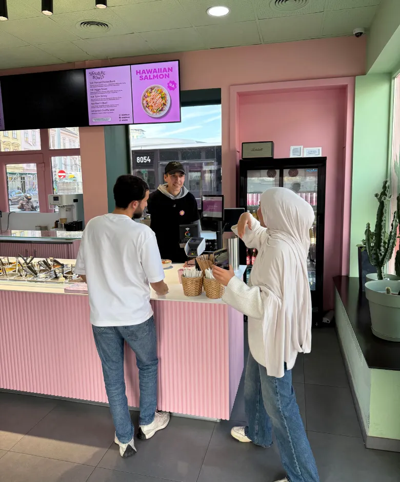 A woman wearing a hijab and a man stand at a pink counter ordering food from a smiling employee in a casual eatery with a menu screen overhead.