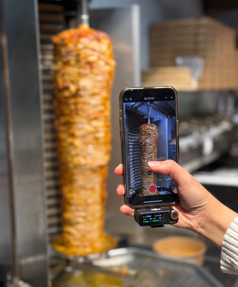 Person holding a smartphone recording vertical rotisserie meat in a kitchen setting.