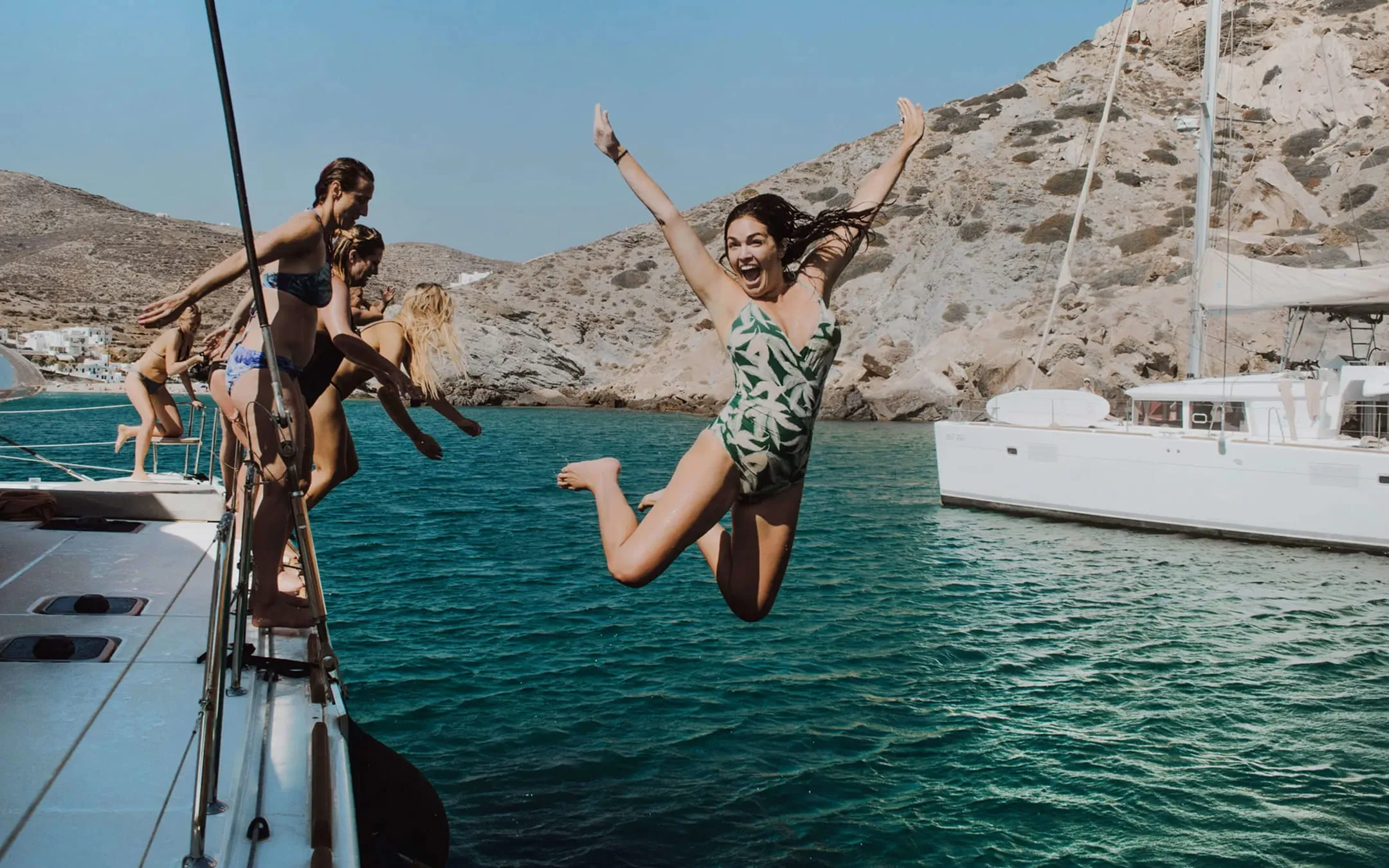 Gruppe von Frauen springt ausgelassen von einem Boot in klares, türkisfarbenes Meer vor zerklüfteten Felsen.