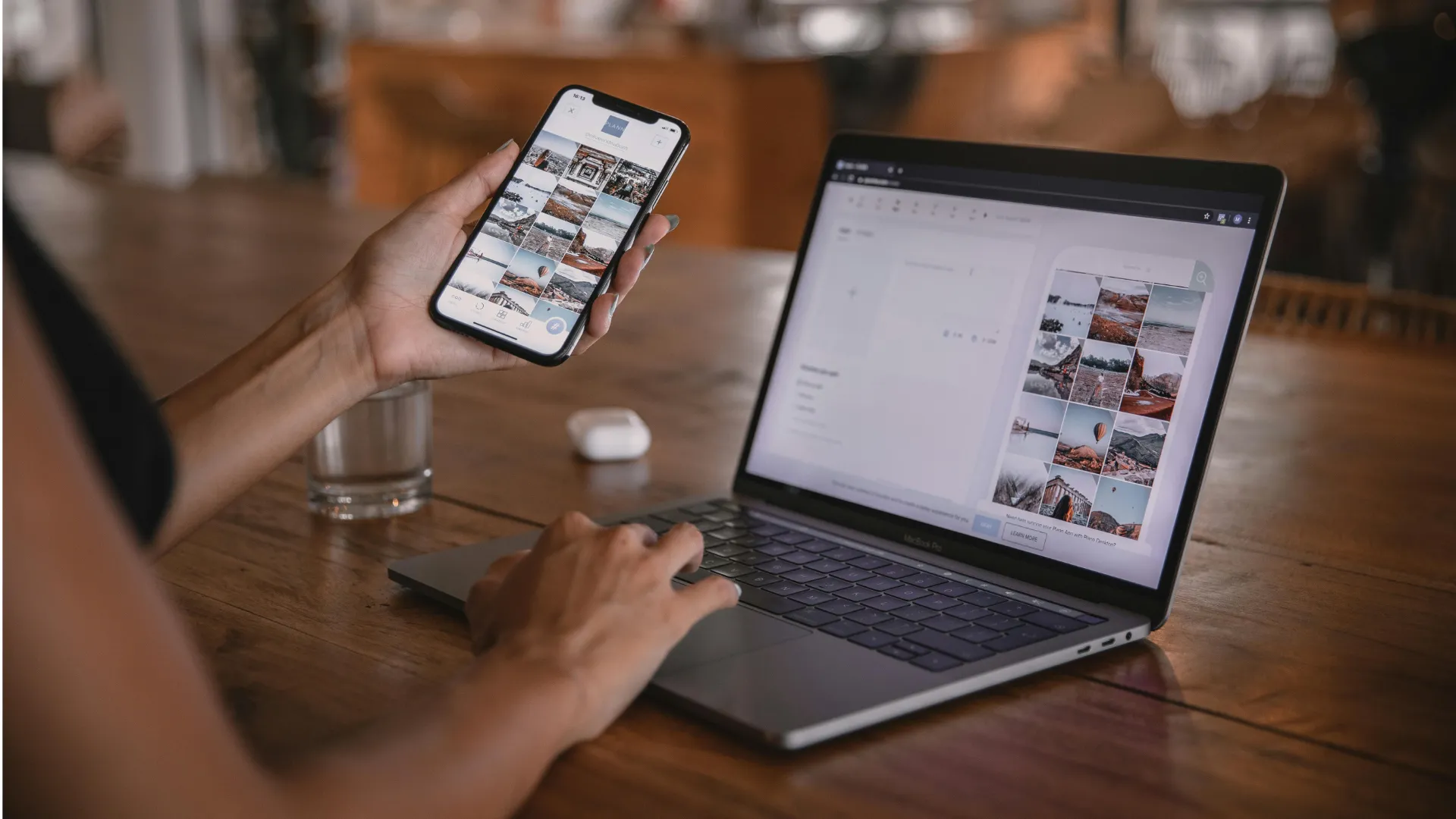 Person hält Smartphone mit Bildergalerie, tippt auf Laptop mit ähnlicher Galerie auf Holzoberfläche.