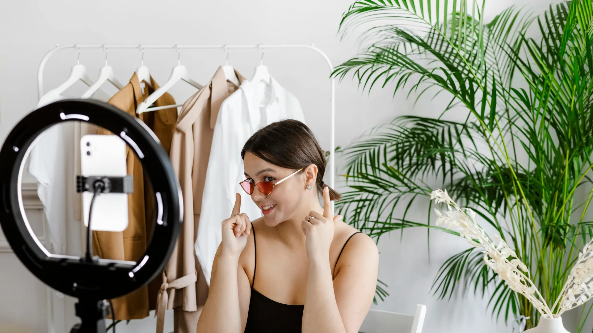 Junge Frau mit Sonnenbrille macht ein Video vor einem Ringlicht mit Kleidern im Hintergrund.