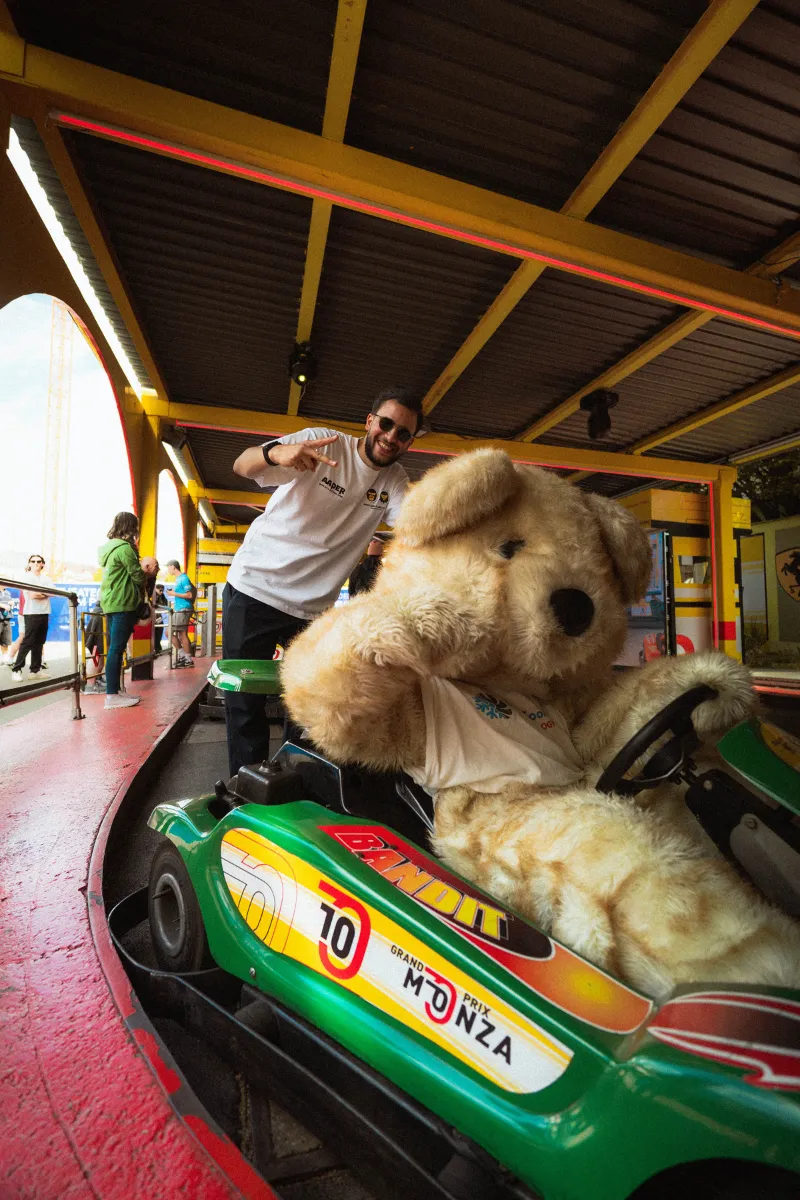 Mann macht Peace-Zeichen hinter großer Teddybär-Kostümfigur in einer grünen Go-Kart-Bahn.