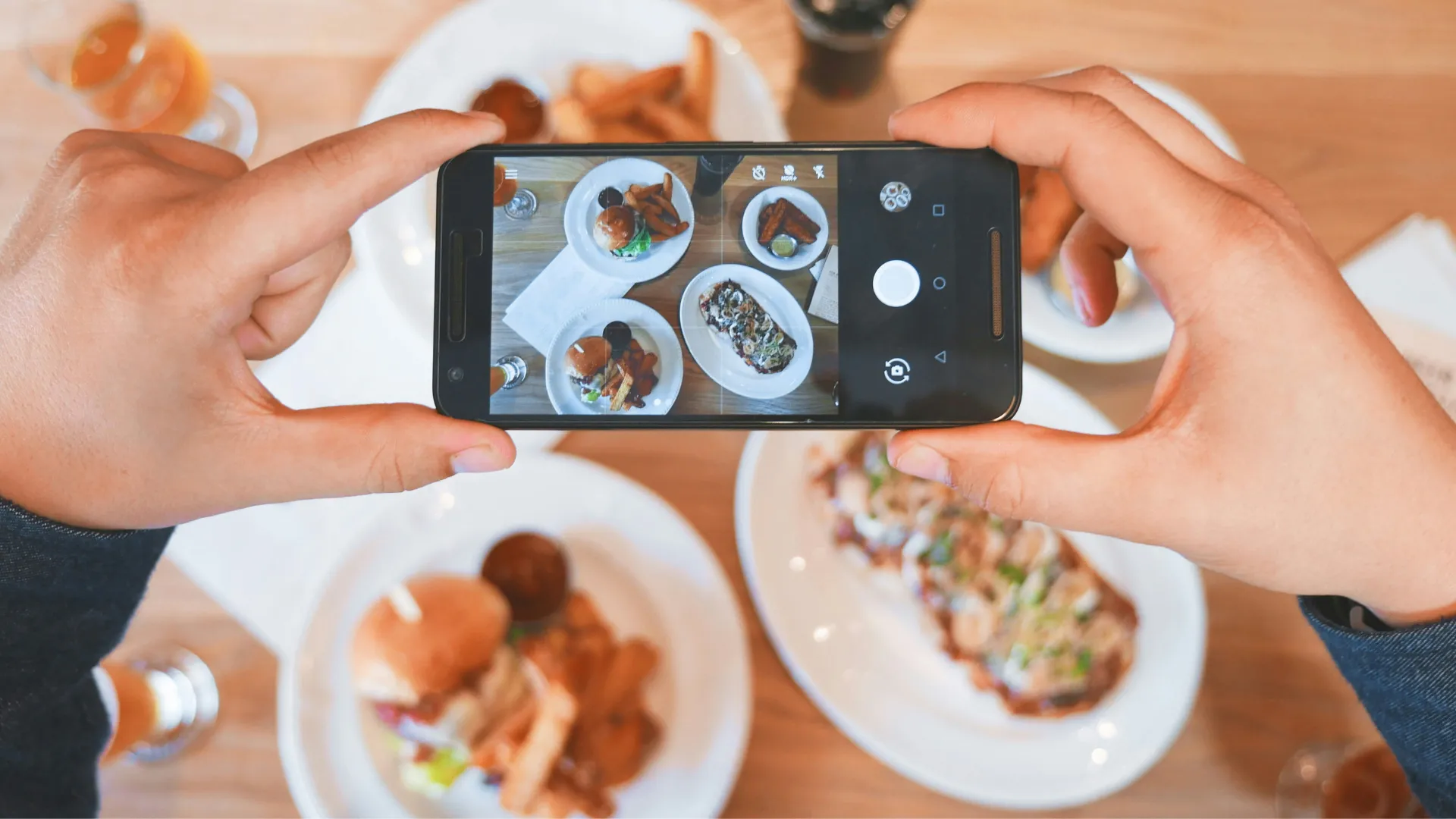 Eine Person fotografiert mit Smartphone servierte Burger, Pommes und ein Gericht auf Tellern.