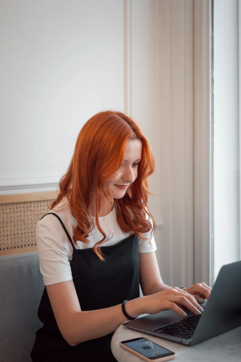 Junge Frau mit roten Haaren tippt auf Laptop, sitzend an einem runden Tisch mit Handy.