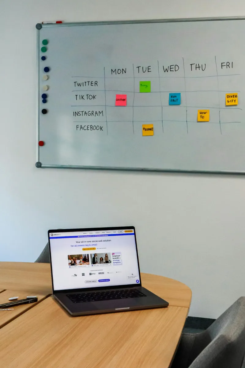 Laptop on a table in front of whiteboard with a weekly social media content plan and colorful sticky notes.