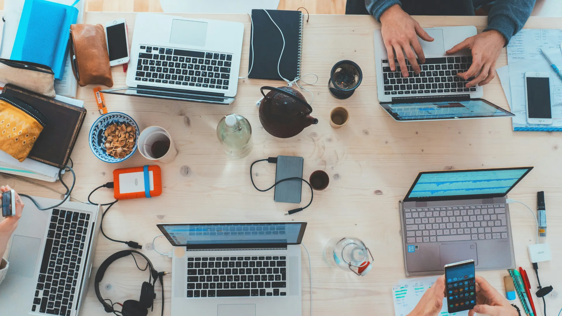 Arbeitsplatz mit vier Laptops, Handys, Notizbüchern, Tasse, Teekanne und Kopfhörern auf Holztisch.