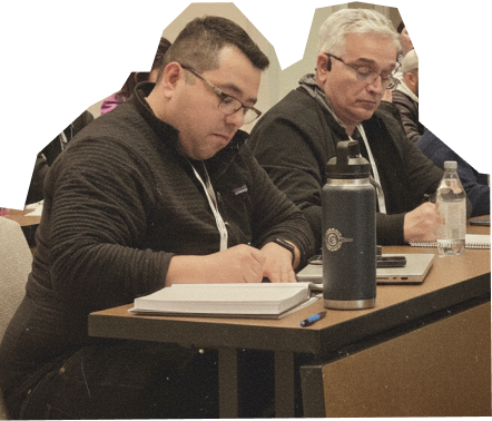 Two attendees at a How to Manage a Small Law Firm event, actively taking notes during a session. One man is wearing a dark sweater and glasses, while the other has light gray hair and a gray jacket. They are seated at a table with notebooks, pens, and water bottles, emphasizing the importance of learning and engagement in law firm management coaching.