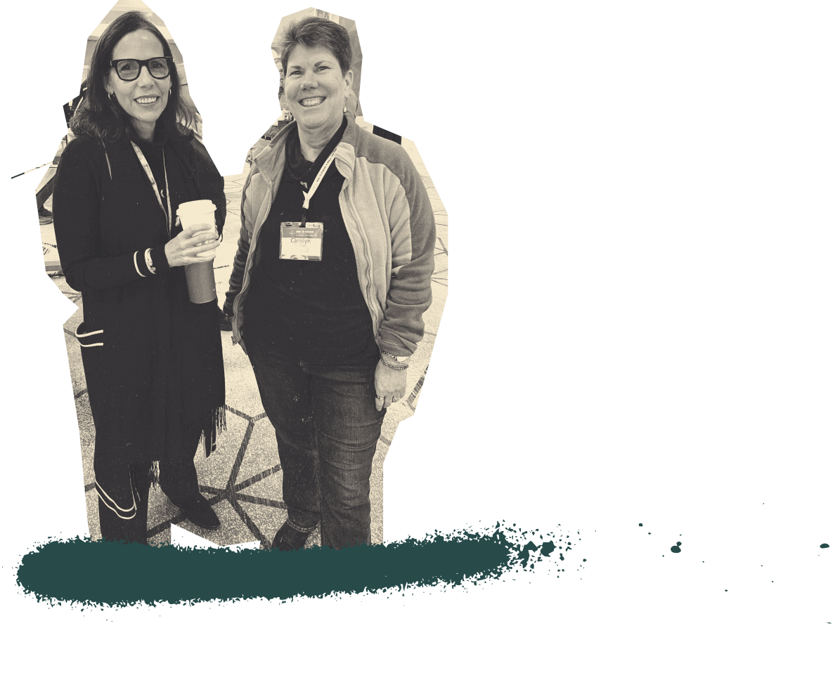 Two women smiling at the camera at a law firm coaching event, holding coffee cups. One woman is wearing glasses, a black scarf, and a conference badge, while the other is dressed in a gray jacket with a conference badge that reads ‘Carolyn.’