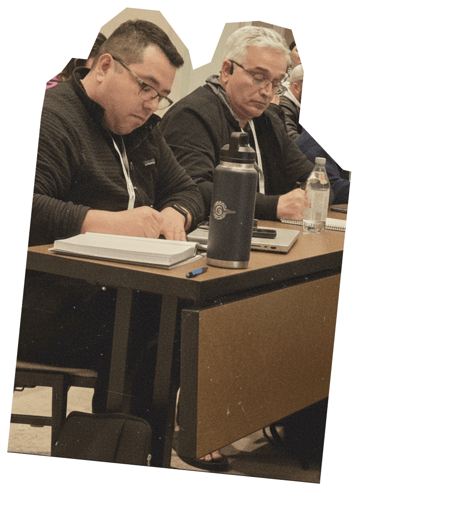 Two attendees at a How to Manage a Small Law Firm event, actively taking notes during a session. One man is wearing a dark sweater and glasses, while the other has light gray hair and a gray jacket. They are seated at a table with notebooks, pens, and water bottles, emphasizing the importance of learning and engagement in law firm management coaching.