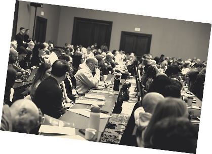 A group of people gathered at a law firm coaching event, engaging in a lively interaction. In the foreground, two women are actively participating in a conversation, with one woman wearing a white sweater and the other dressed in black with red pants. The background shows attendees interacting and networking, with a ‘Resource Center’ sign visible. This image highlights the value of collaboration, coaching, and effective systems in managing a small law firm.