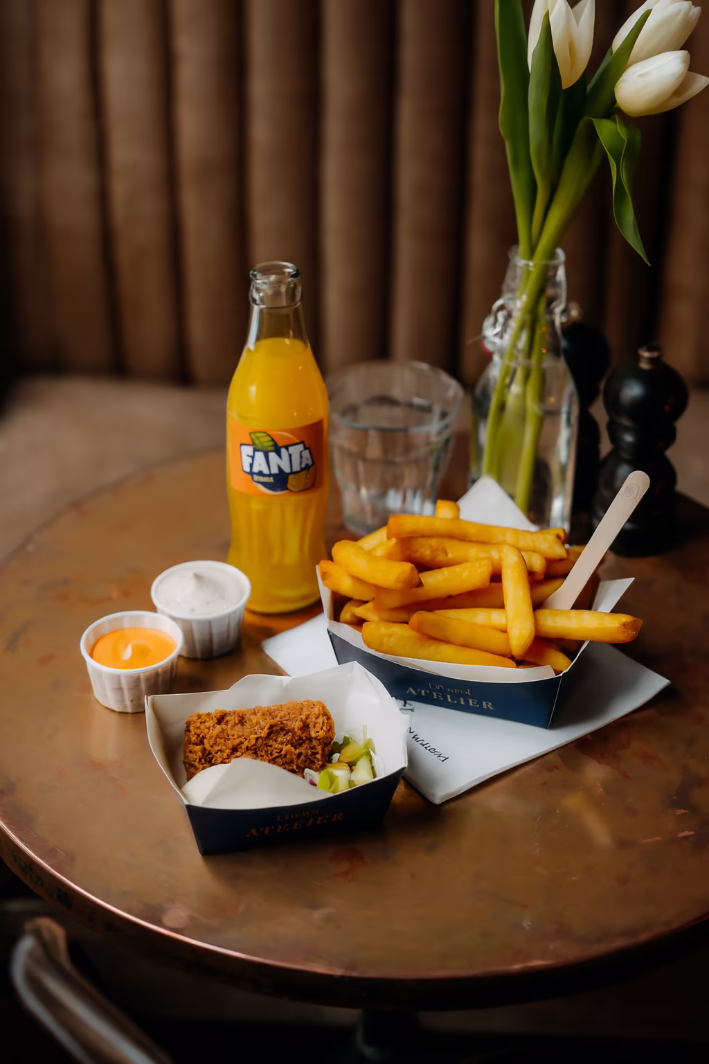 Table with a bottle of Fanta, a glass of water, a serving of French fries with a wooden fork, a piece of fried qroquette with pickles, and two dipping sauces.