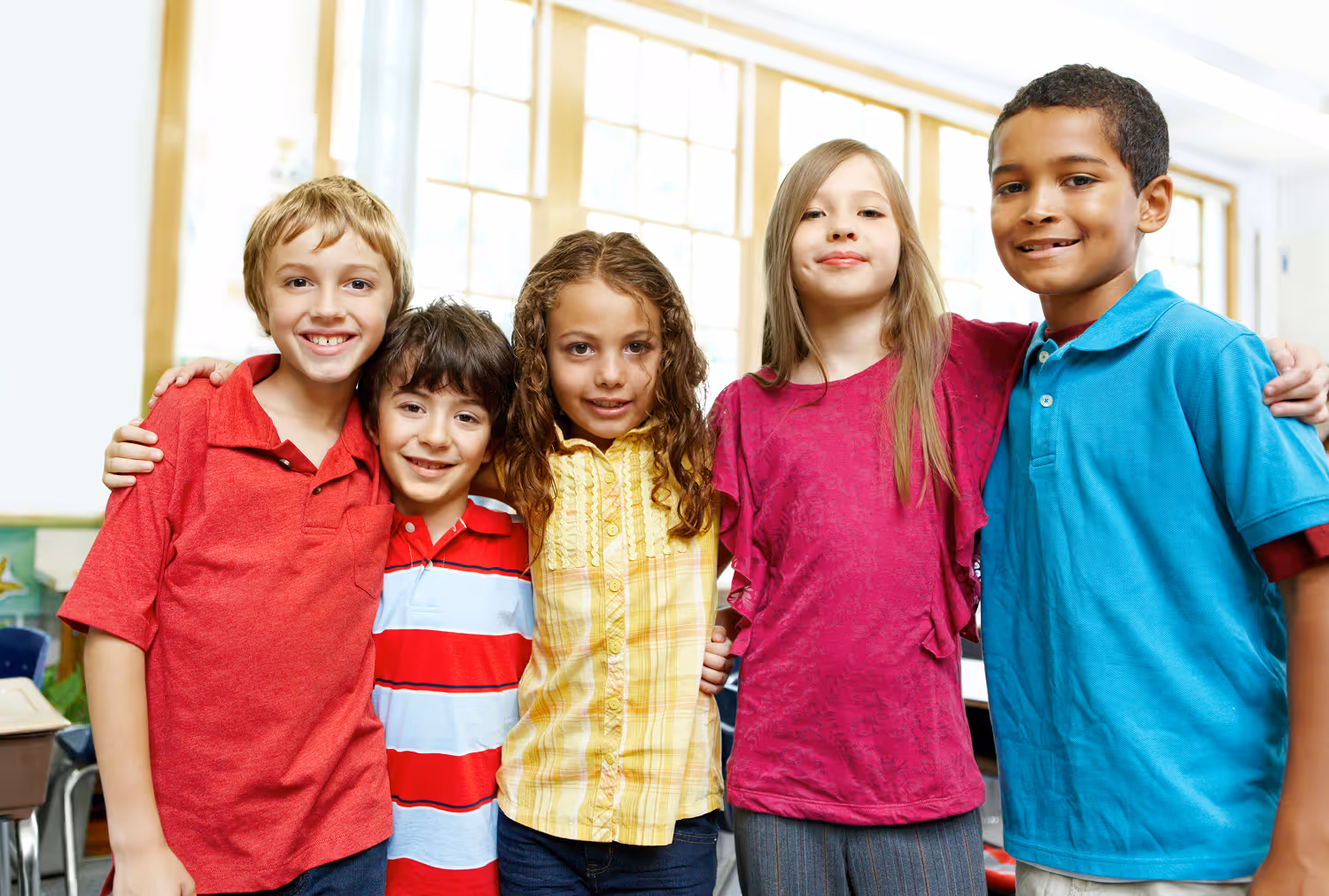 Five smiling children standing close together indoors with arms around each other.