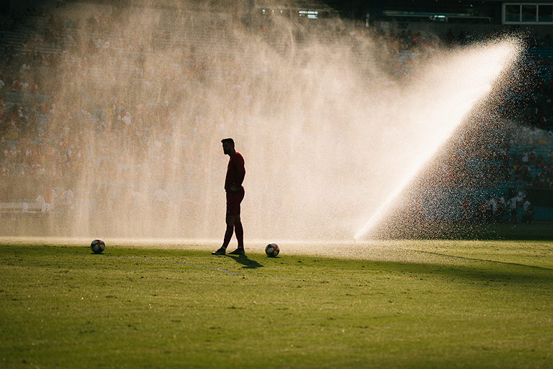 Image of a soccer player standing on a pitch while the sprinkers are going off.