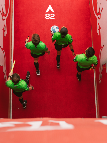 Down shot of four refs walking out of a soccer stadium onto the pitch.