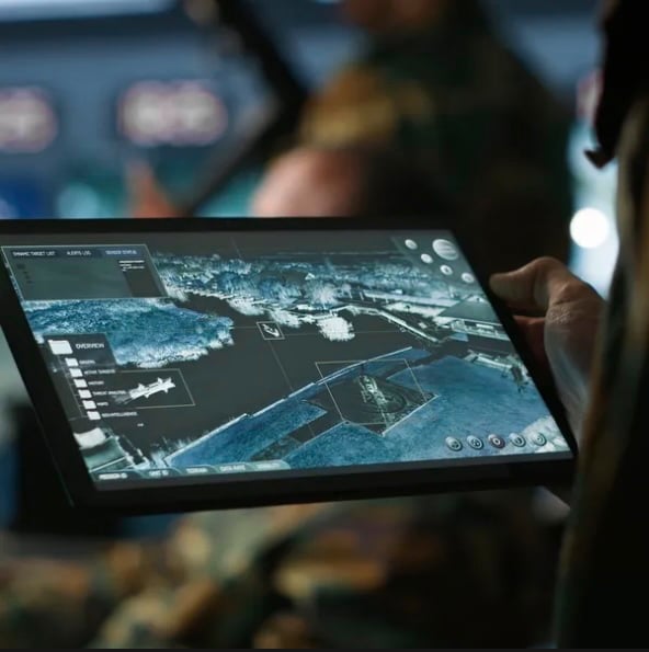 Close-up of a hand holding a tablet displaying a blue tactical map with sensor overlays, with a military operations room blurred in the background.