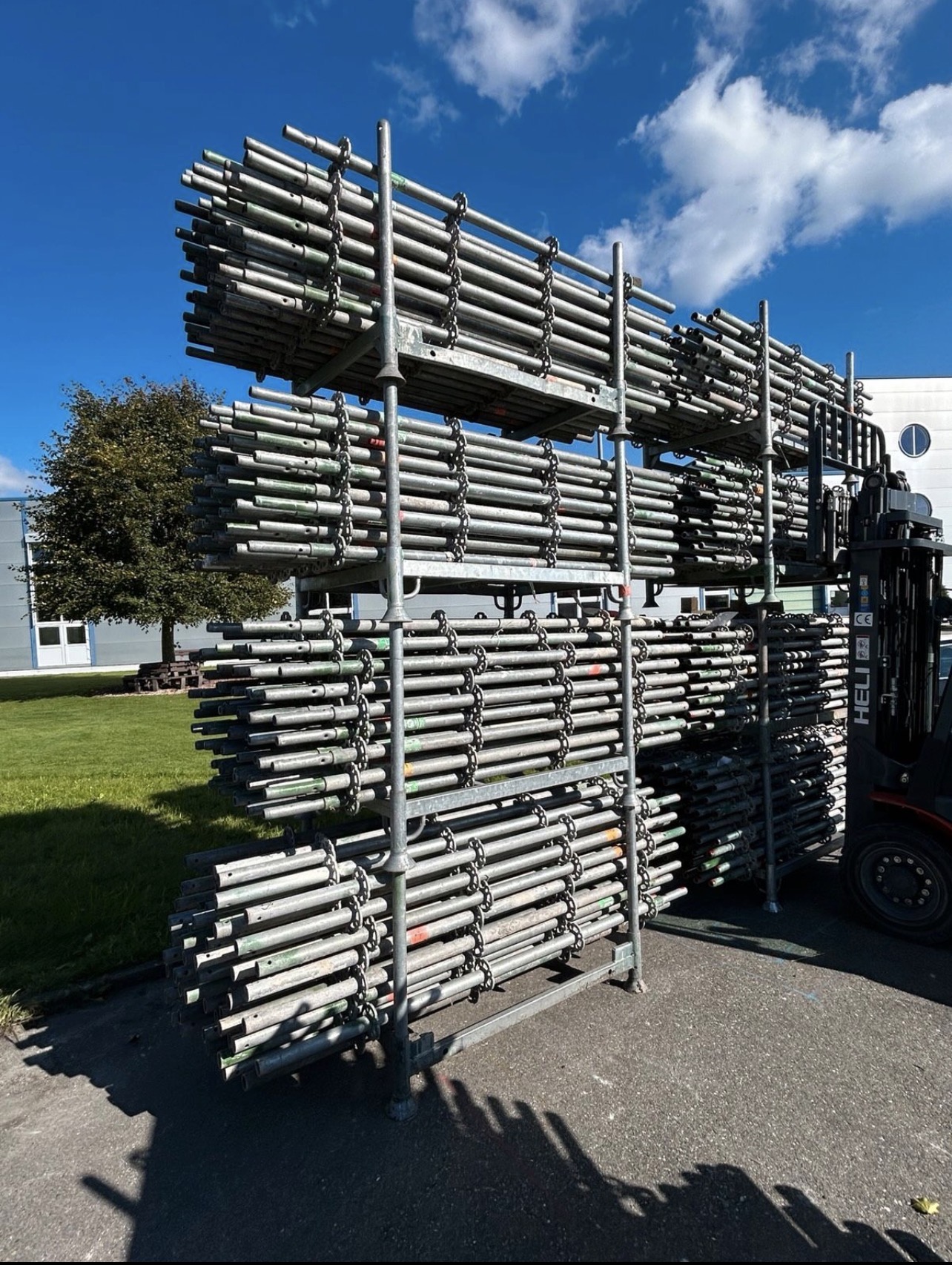 Stacked metal scaffolding pipes on a rack outdoors with a forklift nearby under a partly cloudy blue sky.