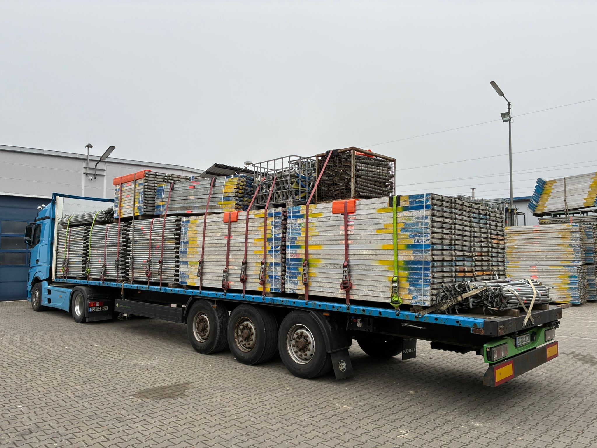 Flatbed truck loaded with stacked metal scaffolding panels secured with colorful straps in an industrial yard.