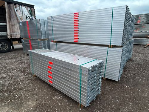 Stacks of silver metal scaffolding planks with green straps and red markings on gravel surface.