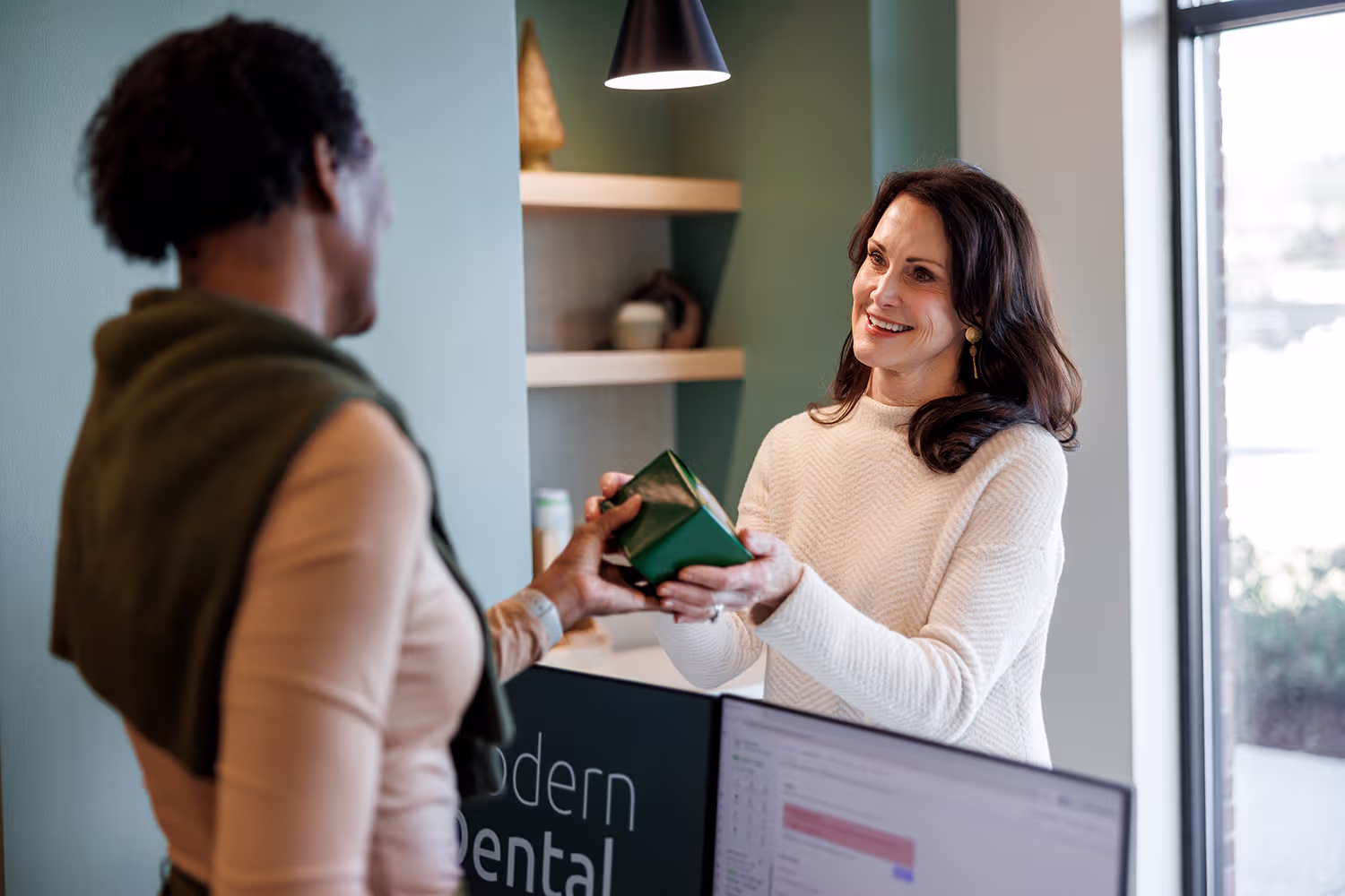 A woman in a white sweater smiling and handing a small green box to another woman standing across a counter in a modern, softly lit office setting.