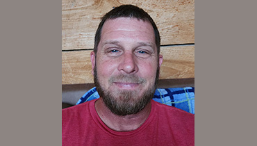 Smiling man with short dark hair and beard wearing a red shirt sitting in front of a wooden wall.