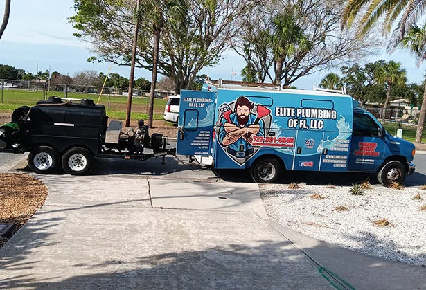 Blue Elite Plumbing of FL, LLC service van with cartoon plumber graphic towing a black equipment trailer on a sunny residential street.