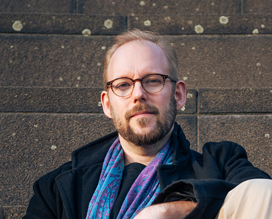 Portrait game designer Iiro Peltonen, a light-skinned man with glasses, a beard, and short hair wearing a black coat and a blue patterned scarf, sitting against a brown textured wall.