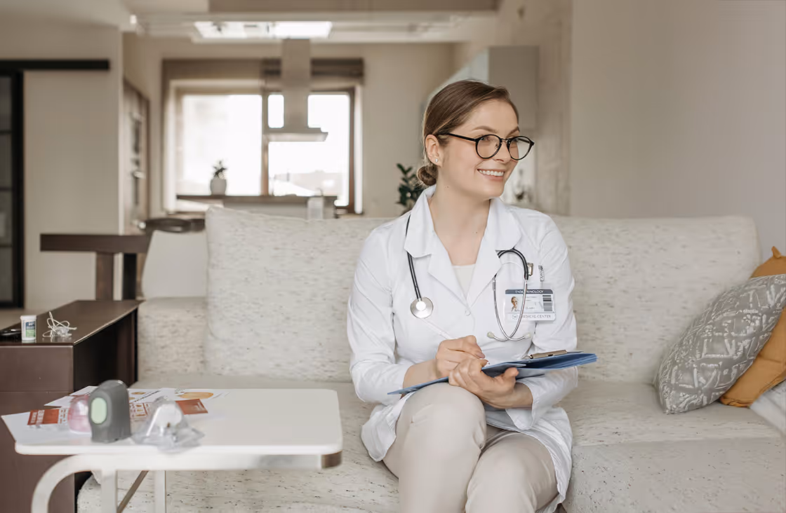 Smiling female doctor in white coat and glasses sitting on a couch holding a clipboard in a modern, light-filled living room.