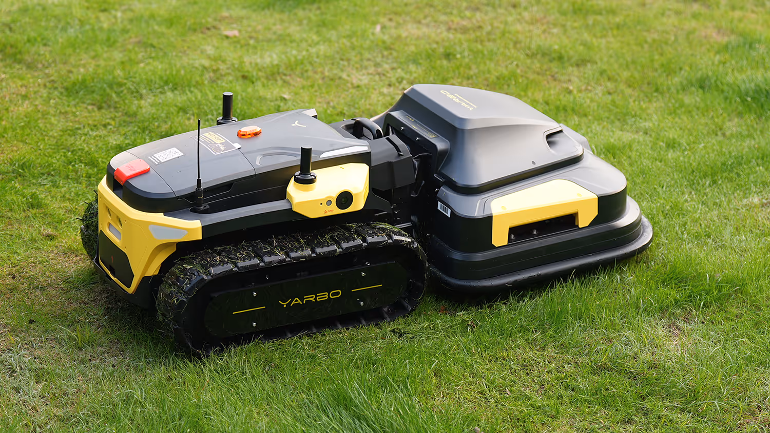 A Yarbo robotic lawn mower with yellow and black tracks cuts grass on a green lawn, featuring a sleek design with a front sensor and antenna.