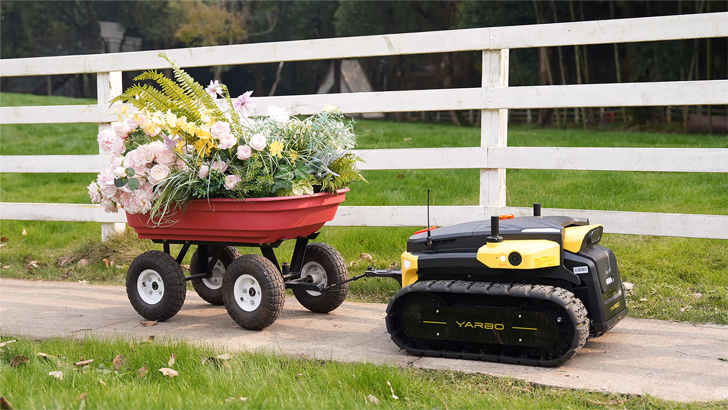 A Yarbo robotic device with yellow and black tracks tows a red cart filled with colorful flowers along a path beside a white fence, set against a green lawn and wooded background.