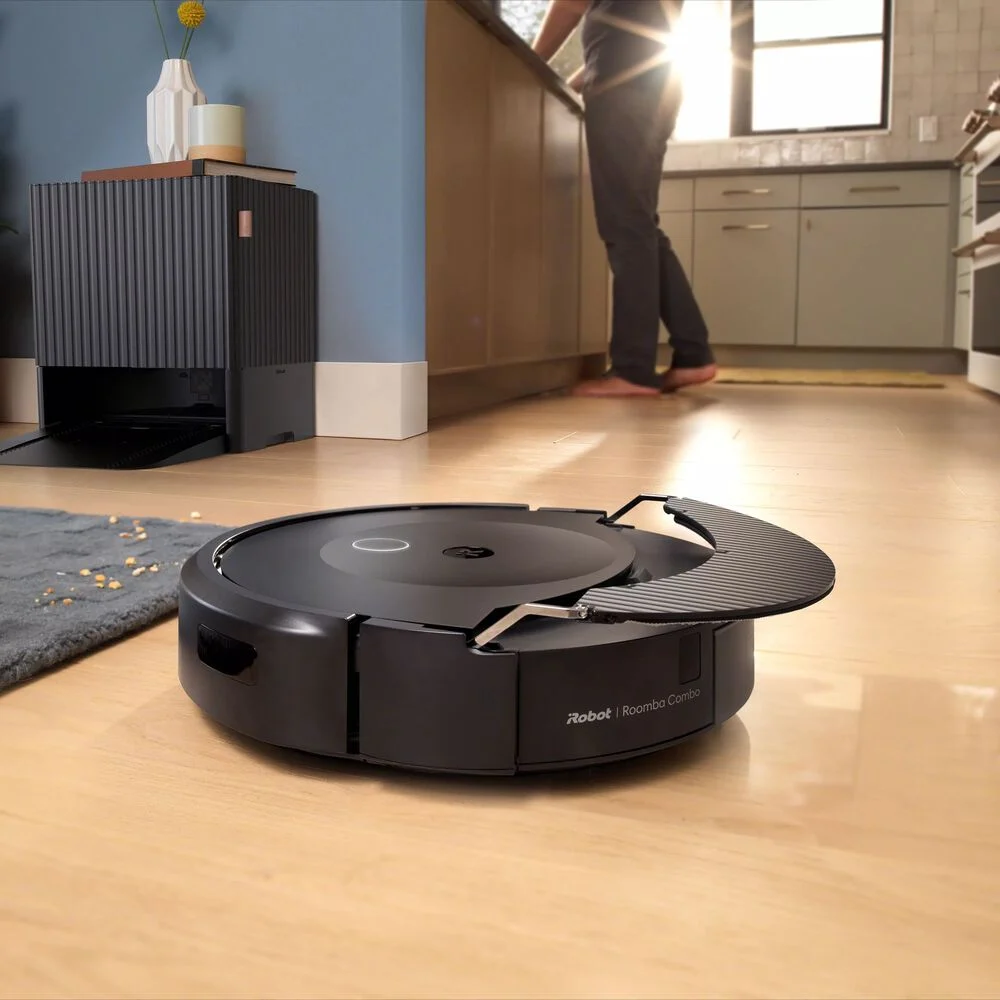 roomba vacum robot riding on wooden flooring with human in background at the kitchen counter