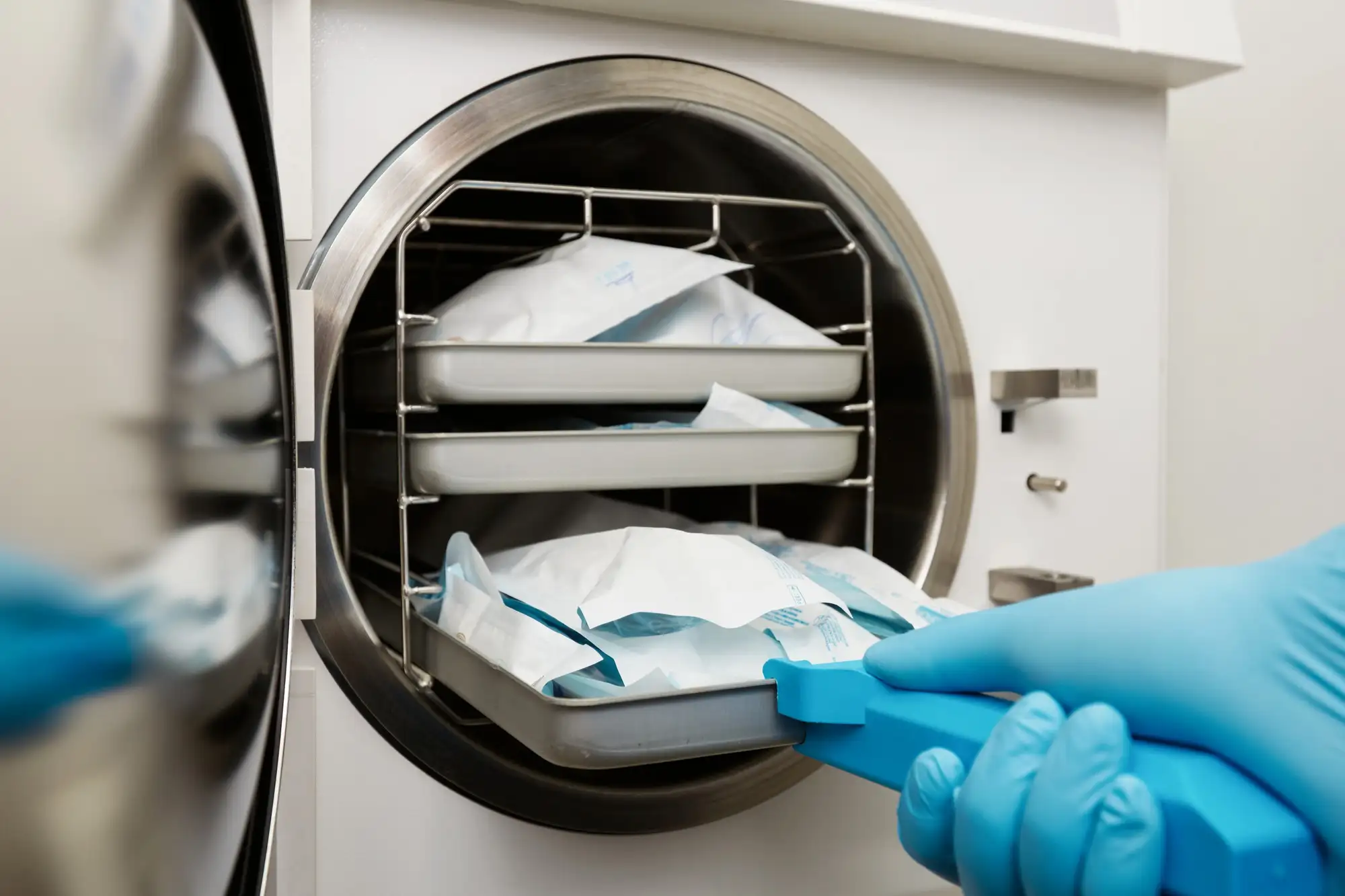 Gloved hand removing a tray of sterilized medical instruments in sealed paper pouches from an autoclave machine.