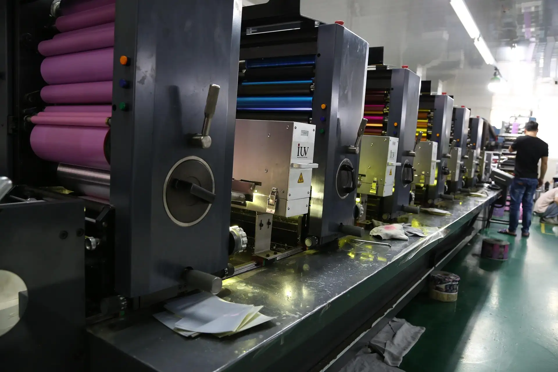 Row of industrial printing machines with colorful rollers in a factory setting and two workers in the background.