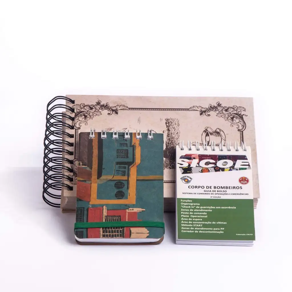 Three spiral-bound notebooks on a white surface, one with vintage decorative cover, one with abstract colorful building design held by a green elastic band, and one small emergency guide titled 'Corpo de Bombeiros'.