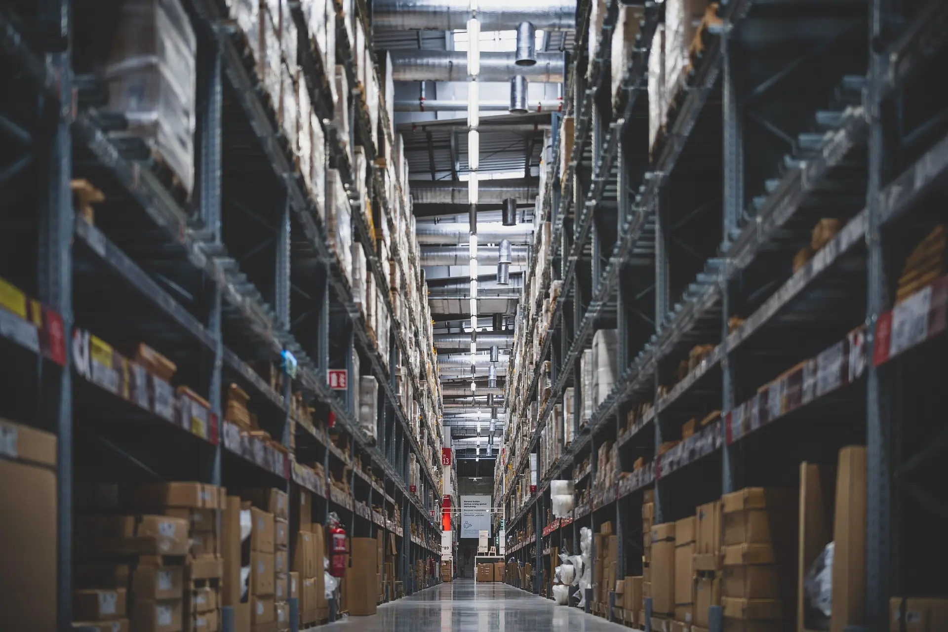 Long aisle in a large warehouse with tall metal shelves stocked with cardboard boxes and wrapped pallets.