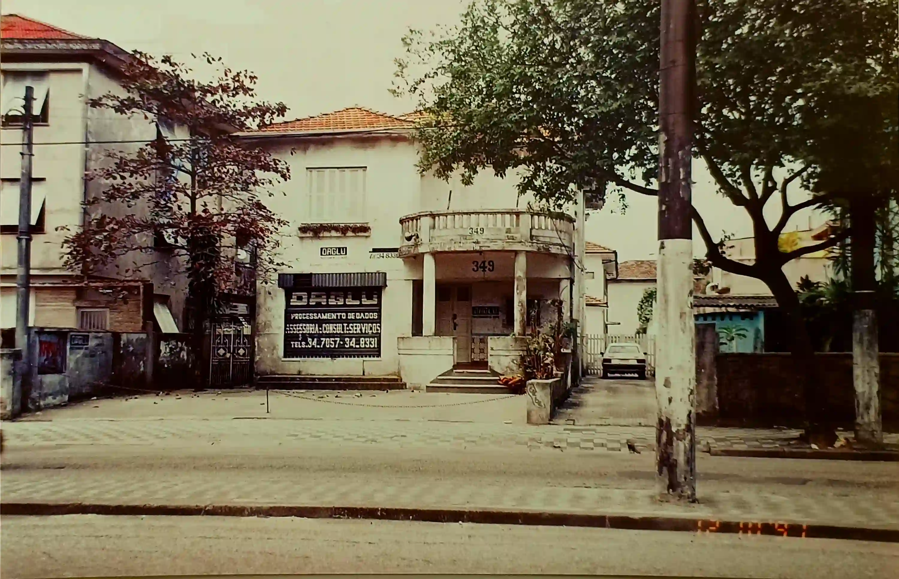 A white two-story building with the number 349 displayed above the entrance and a sign reading 'DAGLU Processamento de Dados' on the left side, with trees and a parked car in the driveway.