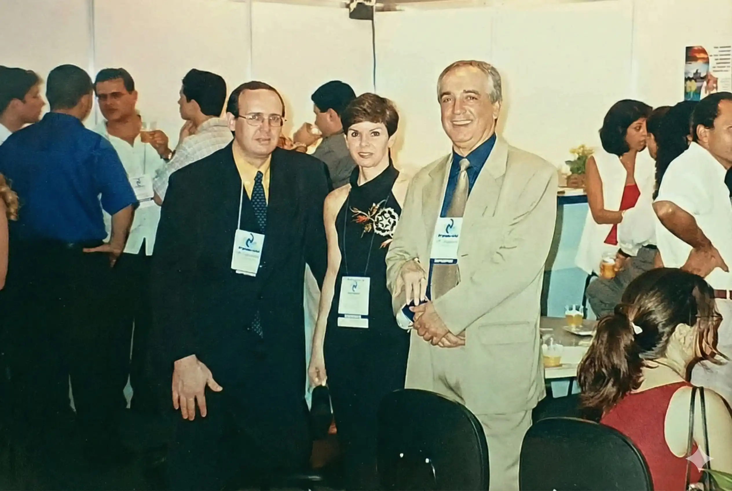 Three people wearing conference badges posing for a photo at an indoor event, with other attendees socializing in the background.