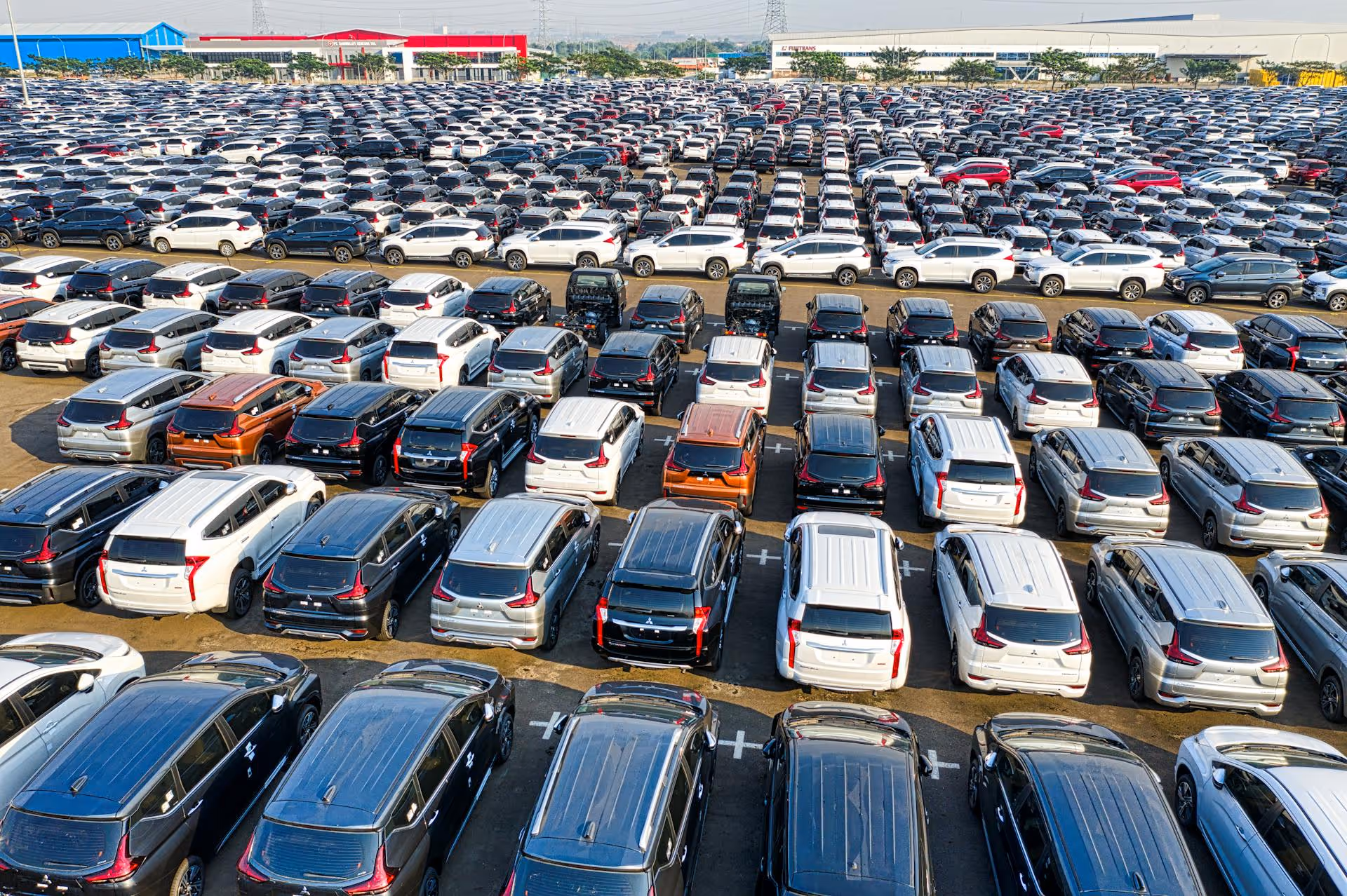 Large outdoor parking lot filled with rows of new black, white, silver, and a few orange SUVs under clear daylight.
