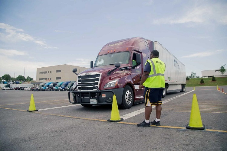 Truck driver in a high-visibility vest guiding a semi-truck through a cone-marked driving practice course in a training lot.