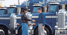 Two men shaking hands beside parked semi-trucks at a commercial truck driving school.