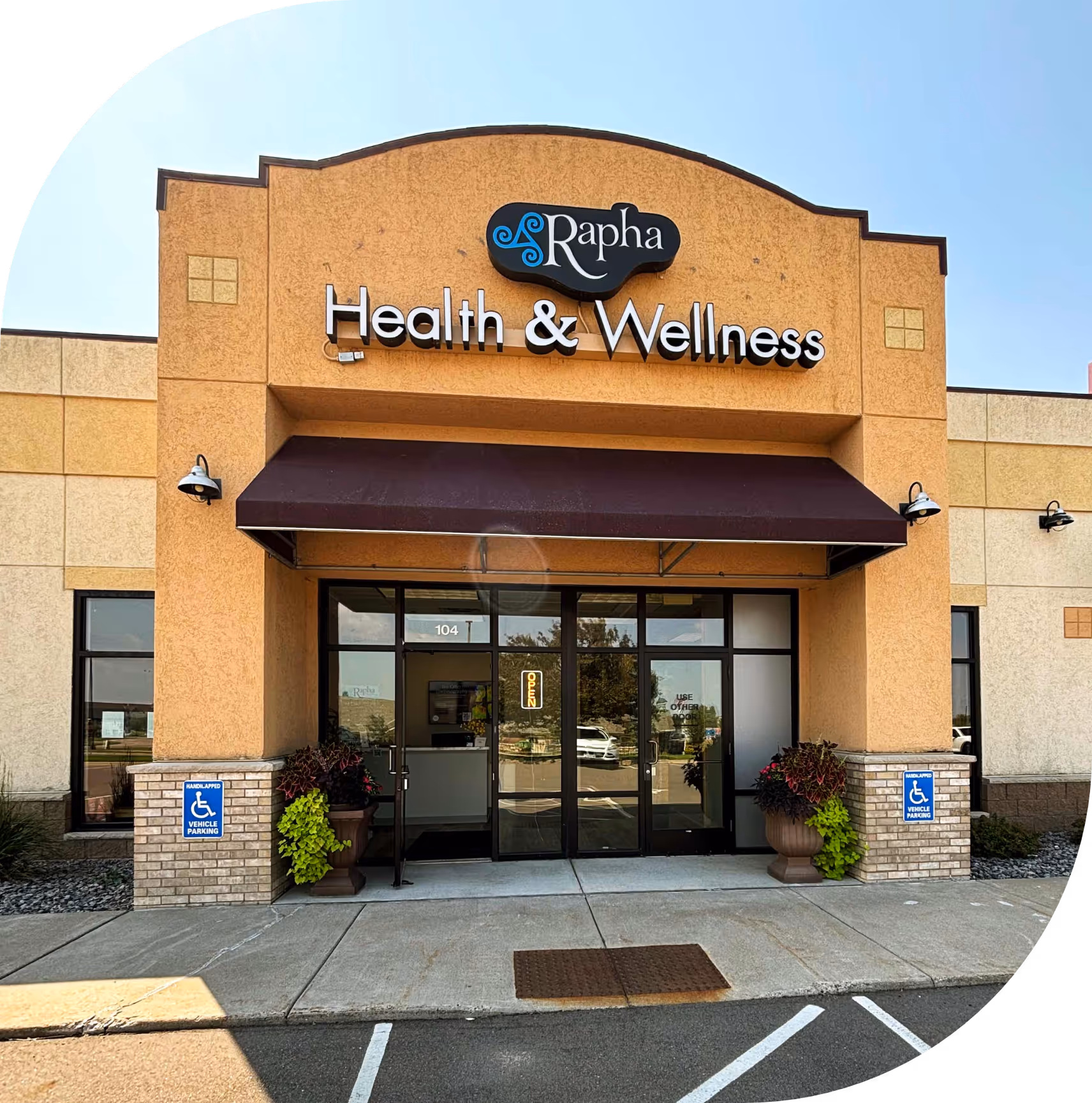 Entrance of Rapha Health & Wellness with double glass doors, wheelchair parking signs, and potted plants.