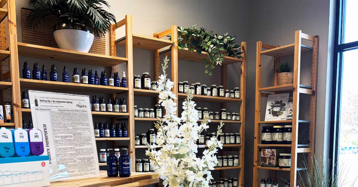 Wooden shelves filled with bottles of supplements and vitamins in a wellness store, with white flowers in the foreground and large window light to the right.