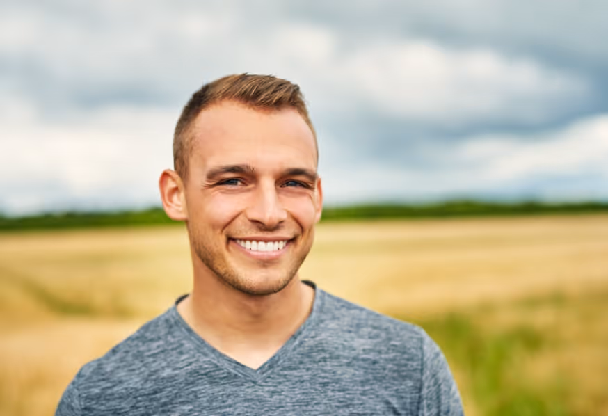 Smiling young man with short hair wearing a gray V-neck shirt standing outdoors in a field under a cloudy sky.