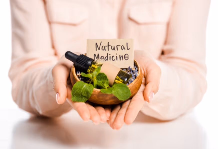 Person holding a small wooden bowl with fresh green leaves, a bottle of oil with dropper, and a sign reading 'Natural Medicine'.