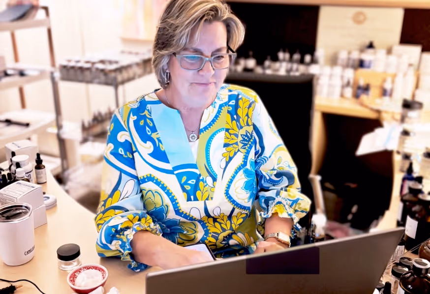 Kimberly Potter wearing glasses and a colorful blue and yellow blouse, working on a laptop at a desk filled with various bottles and containers.