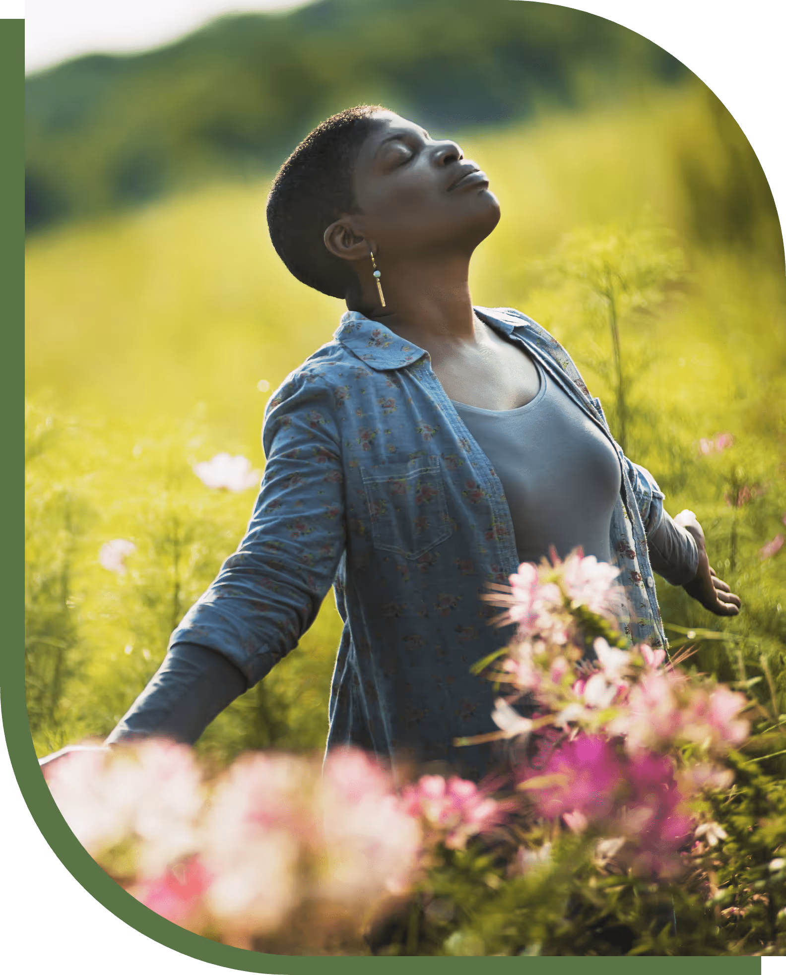 Relaxed woman standing with eyes closed, arms outstretched among pink flowers in a green field.