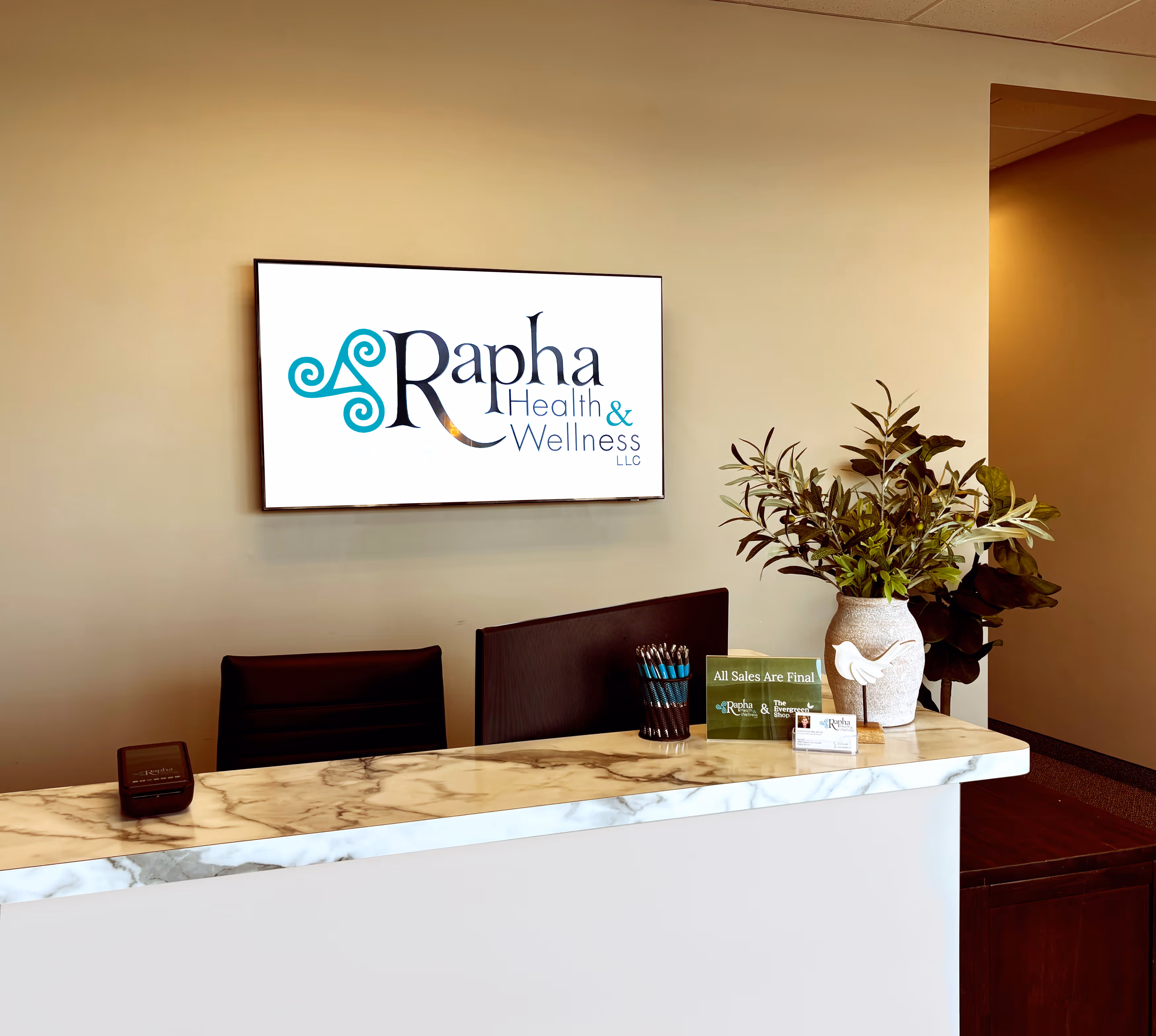 Reception desk with marble countertop, black office chair, potted plant, container of pens, and a screen on the wall displaying Rapha Health & Wellness LLC logo.
