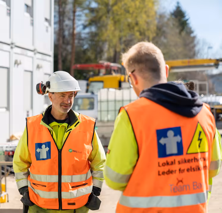 Two construction workers, one wearing a white hard hat and a safety vest, converse at a job site